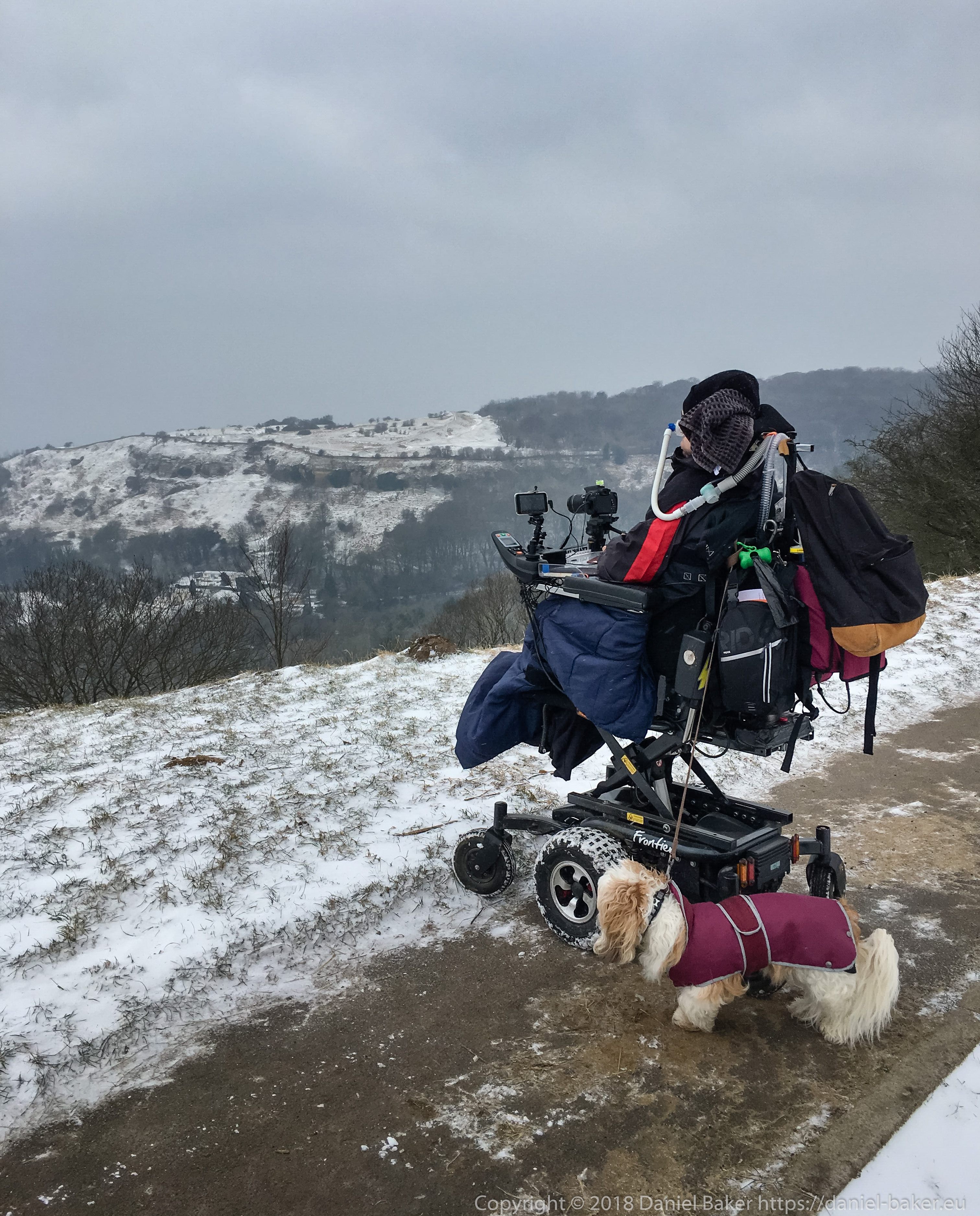 Daniel Baker looking down over Cheltenham from Birdlip hill in the snow, attached to his wheelchair is his faithful dog Mya